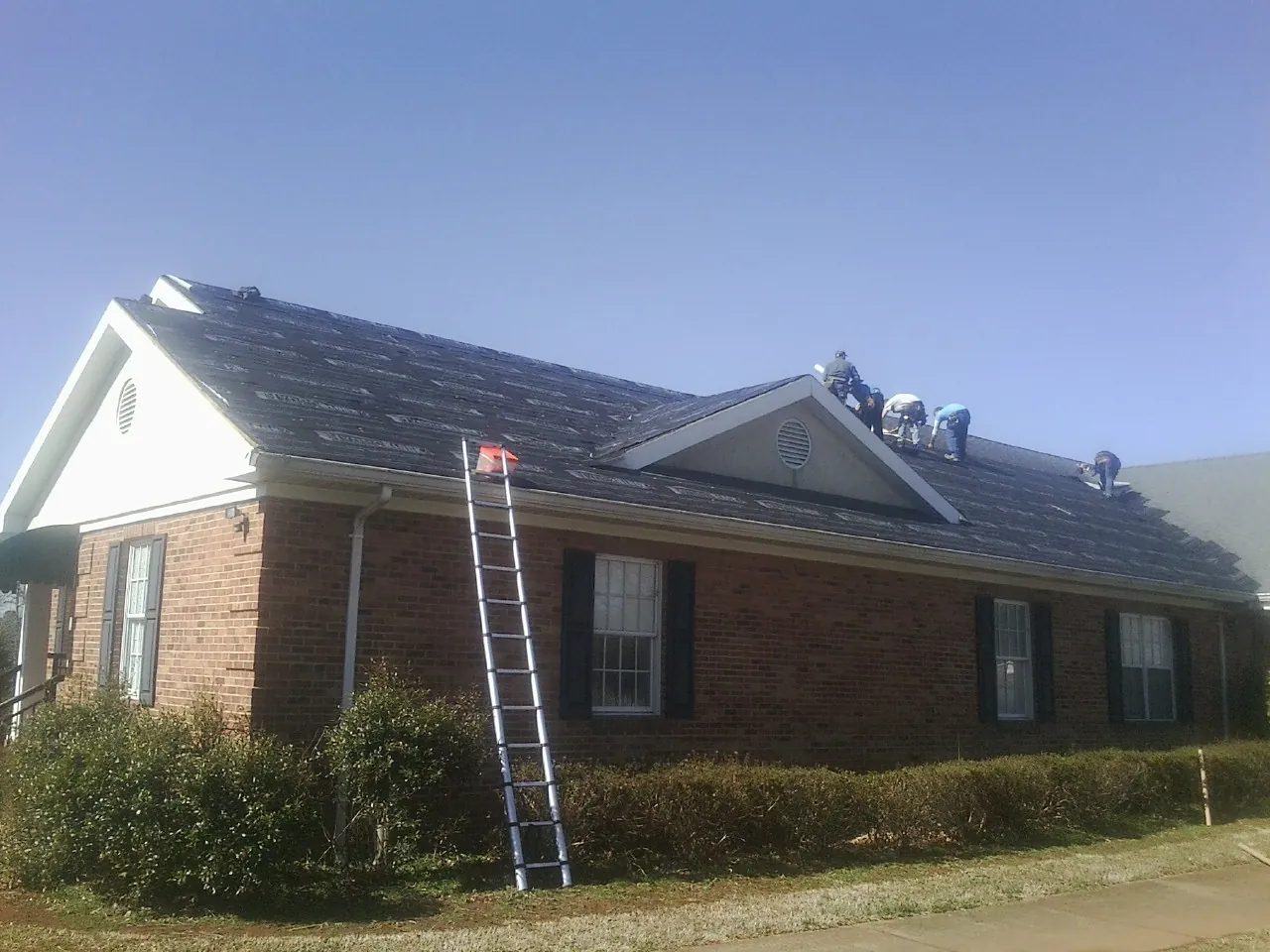 Crew tearing shingles off a brick ranch roof with a ladder set — Emergency Roof Repair work by our crew in Parkers Lake