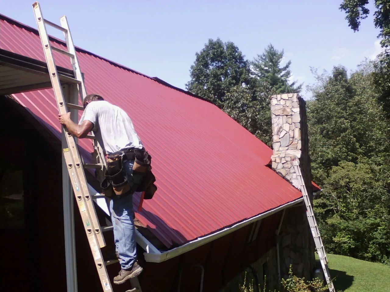 Roofer on a ladder beside a red metal roof and stone chimney — Soffit & Fascia Repair by Roofing Brothers in Parkers Lake